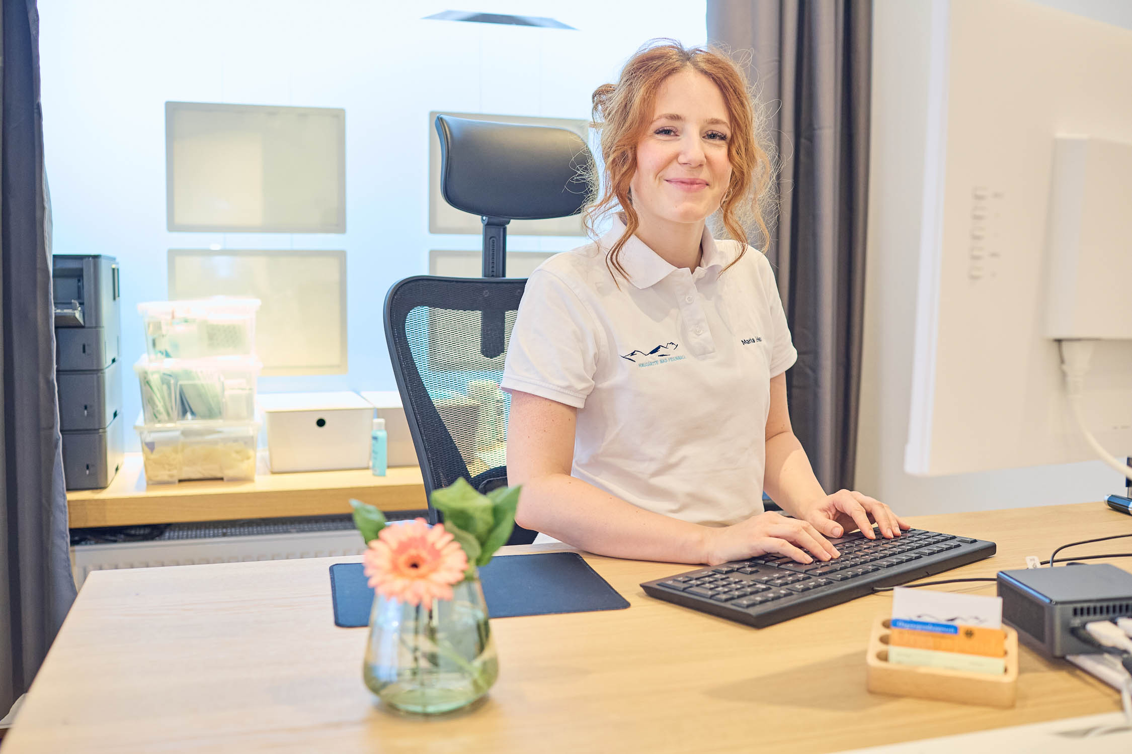 Lächelnde Frau in einem Büro, die an einem Computer arbeitet, mit einem modernen Schreibtisch und Stuhl im Hintergrund.